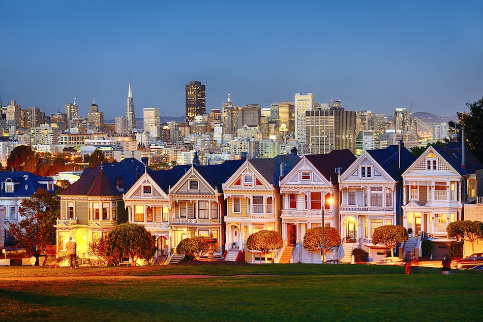 The Painted Ladies of San Francisco, California sit glowing amid the backdrop of a sunset and skyscrapers.